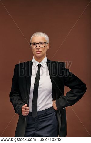 Young stylish manlike woman in elegant black suit and eyeglasses standing in front of camera and looking at you against brown background
