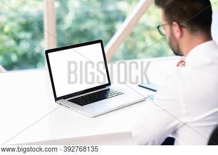 Rear View Shot Of Businessman With His White Fullscreen Laptop Sitting At Office Desk. Focus On Lapt