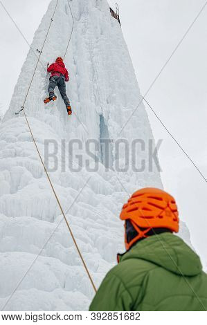 Icebreaker Woman With Ice Tools Axe In Orange Helmet Climbing A Large Wall Of Ice.