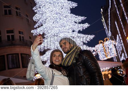 Young couple making selfie outdoors front of huge christmas tree.