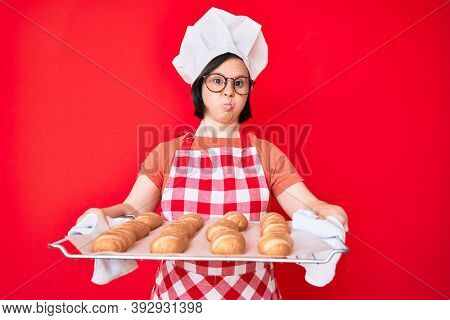 Brunette woman with down syndrome wearing baker uniform holding homemade bread puffing cheeks with funny face. mouth inflated with air, catching air. 