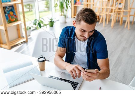 Young designer entrepreneur working on laptop while sittng in his studio work space, using mobile phone