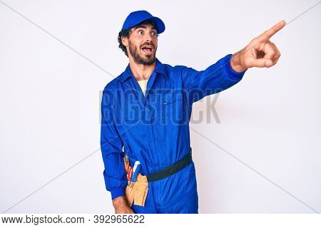 Handsome young man with curly hair and bear weaing handyman uniform pointing with finger surprised ahead, open mouth amazed expression, something on the front 