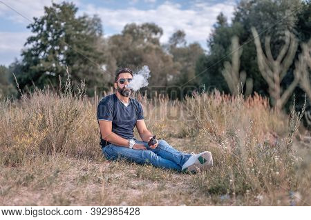 Stylish Brutal Smoker Smoking An Electronic Smoke Device On The Grass. Handsome Man.