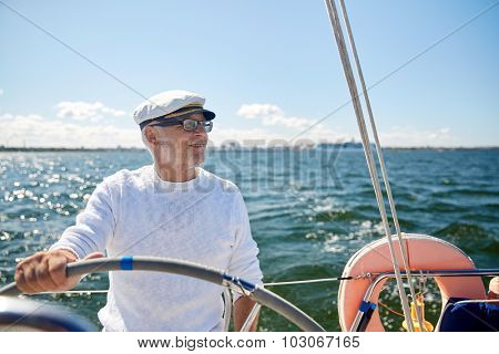 sailing, age, tourism, travel and people concept - happy senior man in captain hat on steering wheel and navigating sail boat or yacht floating in sea