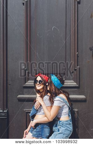 Fashion lifestyle portrait of twins young girls best friends wearing bright make up and similar bandanas smiling and hugging. Have grate time. Urban black old door background.