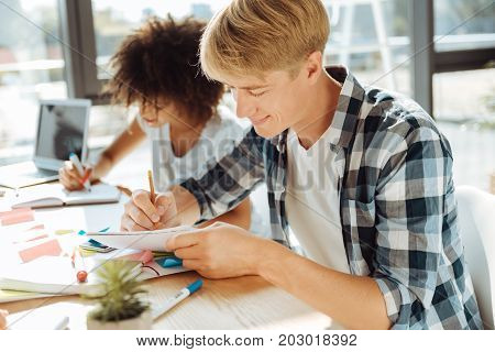 Broaden your mind. Positive male student making notes and sitting at the table while studying with his groupmate