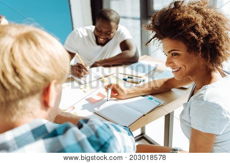 Stay smart. Cheerful young female international student sitting at the table with her groupmates and studying while expressing joy