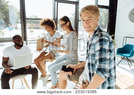 Young and happy. Cheerful handsome young man resting with his friends from university while holding his smart phone