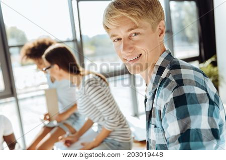 Happy student life. Positive blonde young student smiling while his groupmates sitting in the background while resting after classes