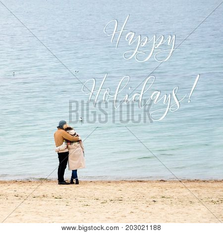 Happy loving couple embracing wearing warm clothes standing on ocean coast. Cold season, sea shore, empty beach in autumn or winter. Happy holidays inscription, greeting card