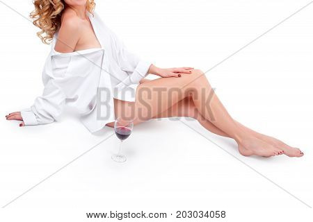Young woman with a glass of red wine on white background. Barefoot curly blonde model dressed in white shirt enjoying wine