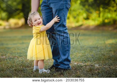 A little girl in a yellow dress hugs her father's pants.