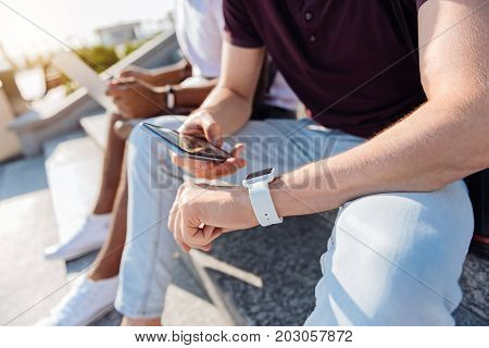 Smart watches. Handsome man putting left arm on his knee and checking time while sitting on the foreground