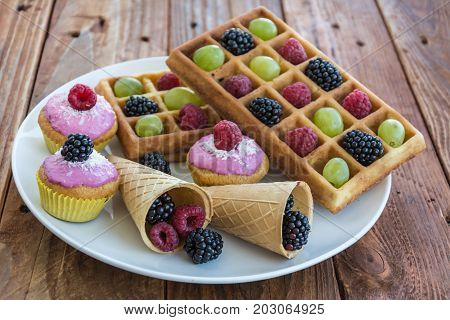 Belgian waffels, cupcakes and ice cream cone with grape, blueberry, raspberry on wooden background
