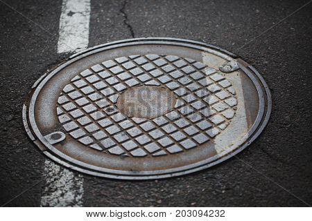 Rustic, metal, vintage, retro, old, cracked hatchway with a geometrical ornament on a gray pavement road background. Dirty hatchway on the road surface closing canalization with sewerage.