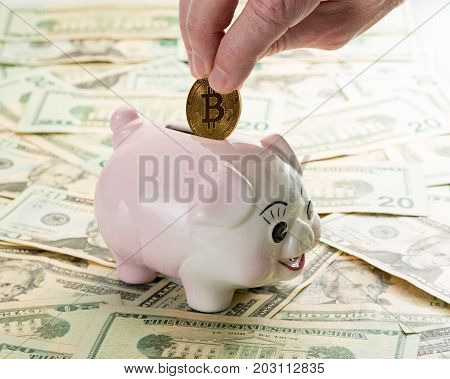 Fingers placing a single gold bitcoin coin into the slot on a piggy bank with 20 dollar notes on table