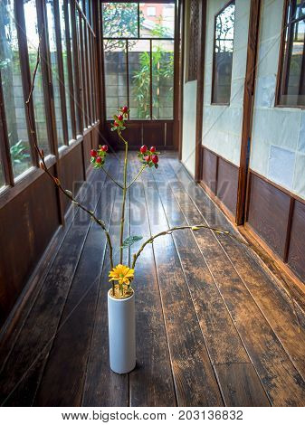 Close up of a flower vase with a yellow flower and red branch at indoor view of hall with a wooden floor, in Kyoto, Japan.