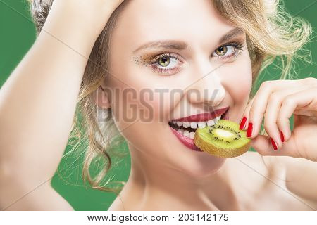 Closeup Portrait of Sensual Caucasian Blond Model With Disheveled Hair Biting Slice of Green Juicy Kiwi Fruit.Against Green Background. 