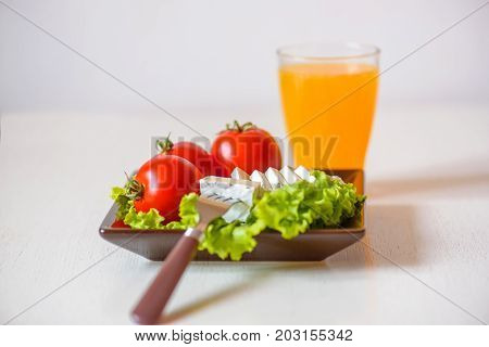 Tomatoes and cheese slices lie on a sheet of fresh salad in a plate. A glass of orange juice stands side by side on the table.