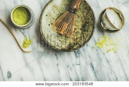 Flat-lay of Japanese tools and bowls for brewing matcha green tea. Matcha powder in tin can, Chashaku spoon, Chasen bamboo whisk, Chawan bowl, cups, grey marble background, top view, copy space