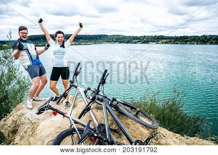 cuple of sportmen sitting near lake with bicycles and showing hands up
