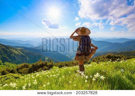 On the lawn in mountains landscapes the hipster girl in dress stockings and straw hat stays watching the sky with clouds.