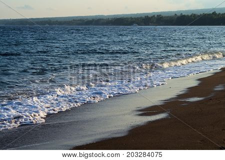 Blue sea water over volcanic sand beach. Relaxing sea wave surf over seashore. Coastal sunset. Idyllic seascape for summer vacation. Exotic island seaside banner. Sunny day on beach. Seawater foam