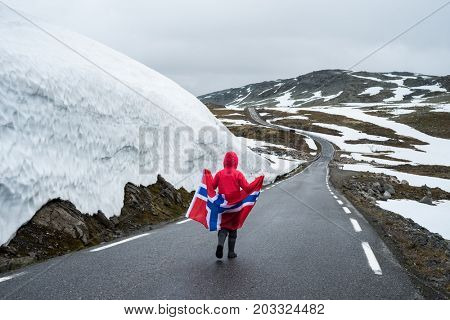 Bjorgavegen - Snowy road in Norway. Girl with the Norwegian flag on a mountain road near a wall of snow. Tourist Route Aurlandsfjellet runs from Aurlandsvangen to Laerdalsoyri