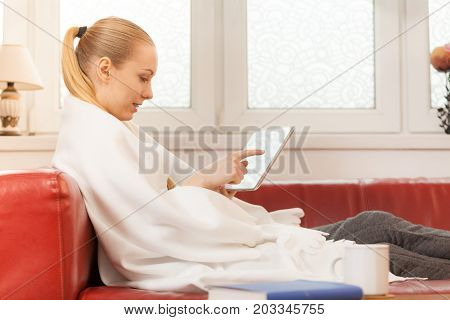 Young woman lying on sofa under warm blanket relaxing using tablet reading news and checking social media having fun during leisure time. Modern relaxation.