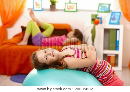 Laughing schoolgirl lying on gym ball, looking at camera, friend lying on sofa in background.?