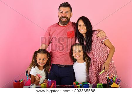 Girls man and woman with happy faces by their art desk. Parents and children paint with markers and smile on pink background. Artists family hugs and draws. Family leisure time and art concept