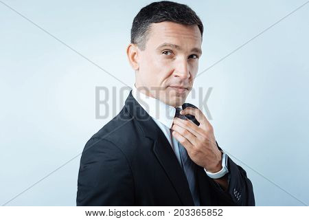 Office style. Joyful nice confident man wearing a suit and fixing his tie while getting ready to work