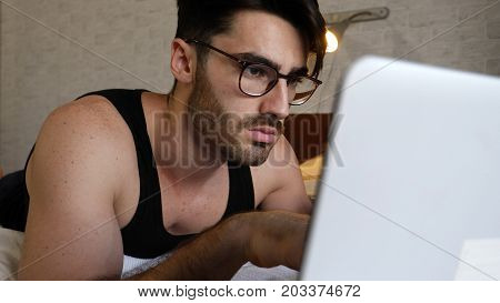 Attractive Young Man with Serious Expression, with Laptop on Bed Working on his Start-up Business - Young Male College or University Student Doing Homework, in Bedroom
