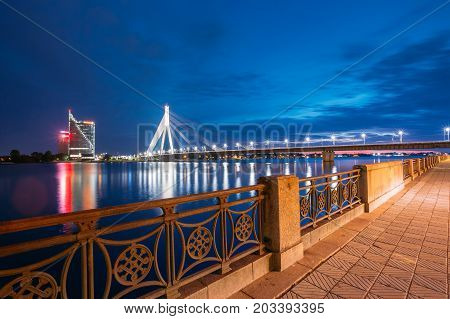 Riga, Latvia - July 01, 2016: View Of Famous Vansu Cable-Stayed Bridge In Bright Night Illumination From The Deserted Embankment Of The Daugava Or Western Dvina River Under Dramatic Blue Summer Sky.