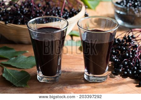Two glasses of elderberry syrup on a wooden table with fresh elderberries in the background