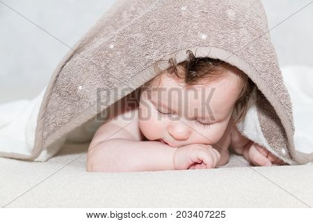 Close up portrait of cute three month old baby girl covered with a bath terry towel over her head lying on tummy in the bathroom. Little newborn girl lying on a white cover. Sleeping kid after showering.
