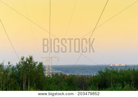 The tip end anchor metal supports of overhead power lines with wires is on the slope of the hill above the forest trees on a background of soft orange colorful sunset and the city buildings