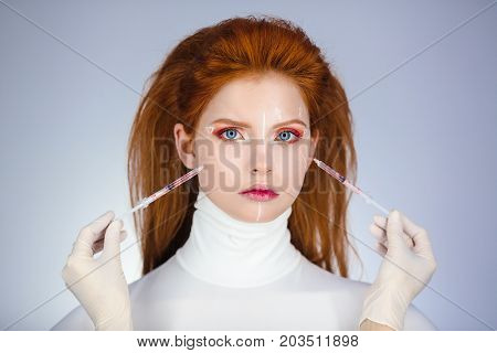 Beautiful Woman before Plastic Surgery Operation Cosmetology. Beauty Portrait Fashion Concept. Treatment with botox or hyaluronic collagen injection. Young red hair girl holding syringes in hands with correction mark for plastic surgery on her sad face wi