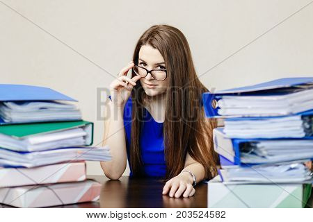 Young Beautiful Girl In Glasses And Blue Dress Is Sitting In The Office At The Table With A Lot Of F