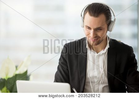 Smiling businessman in headphones looking at laptop screen at workplace. Friendly young manager participating in online meeting or conference, remote job interview, learning foreign languages.