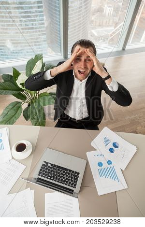 Distraught businessman at work looking up screaming in anger, with report documents, laptop, coffee on desk. CEO deranged because of financial failure. Heavy workload, profit loss, stock market crash.