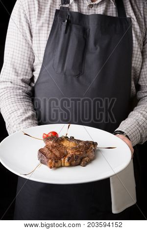 Waiter offering delicious restaurant dish. Rib eye steak decorated with tomato in unrecognizable male hands. Copy space on dark background.