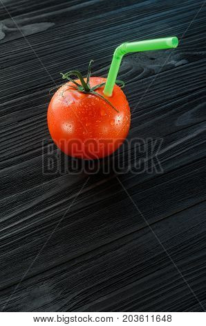 One red tomato with straw on black wood background. Fresh juice healthy food concept. Tomato as a vessel with fresh juice. Background with a Blank Space for Text