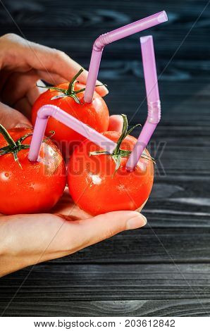 Three Ripe Red Tomatoes with Straws in Hands on Black Wooden Background. Fresh Juice Healthy Food Concept. Tomato as a Vessel with Fresh Juice