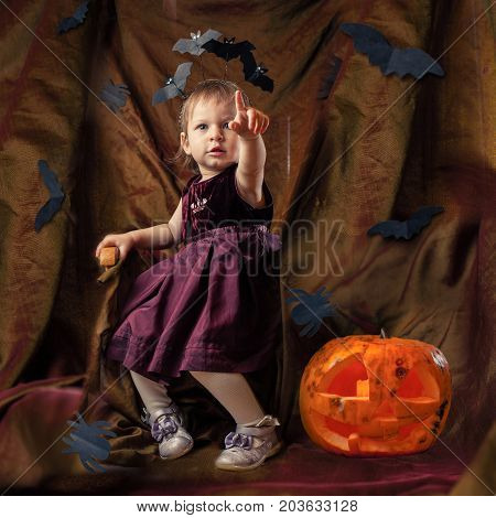 Decorations for Halloween pumpkin carved with scary eyes - lights, bats flying around. The child sits on the throne, and his finger pointing to something ahead. Staging decorative, entertainment for the holiday.
