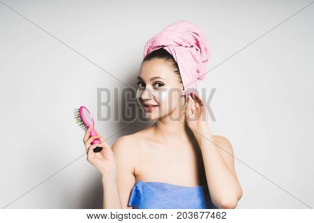 A joyful girl after bathing with a towel on her head is holding a pink comb in her hands, gazing at the camera