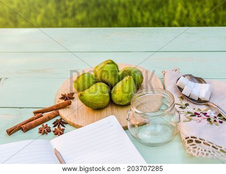 Still life with figs, jam cooking process. Fresh green figs on wooden backround, surrounded by spices, embroidered nupkin, vintage spoon, a glass container for jelly. Notebook with copy space.
