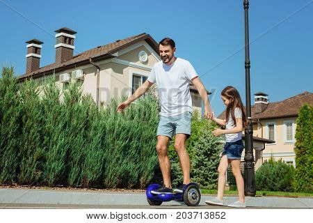 Always supporting. Charming little girl holding the hand of her father and providing support to him while the man riding a self-balancing scooter