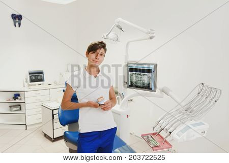 Woman Dentist With Short Hair In A White Shirt And Blue Trousers Holds Dentures In The Dental Office